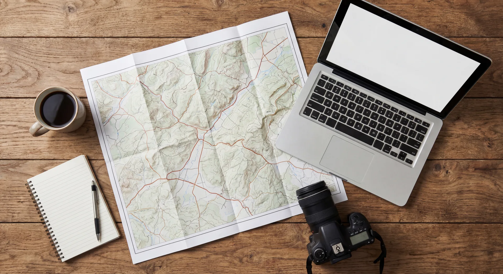 Flat lay of RV trip planning items including map, laptop, coffee, and notebook on wooden table