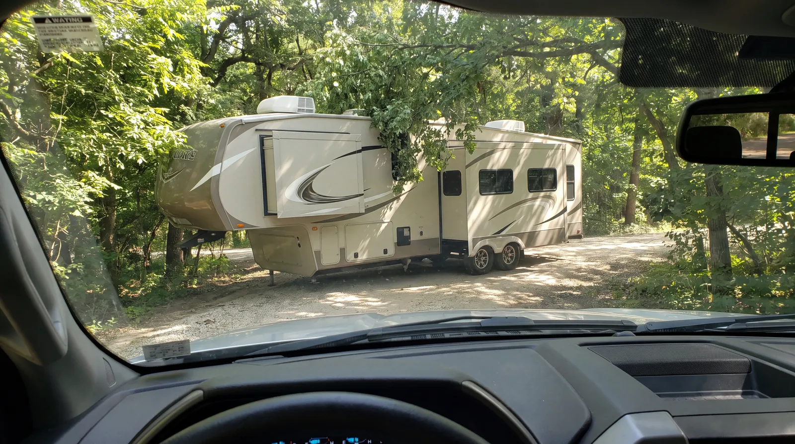 Driver view of a long fifth wheel RV turning into a narrow wooded campground loop entrance with branches close overhead