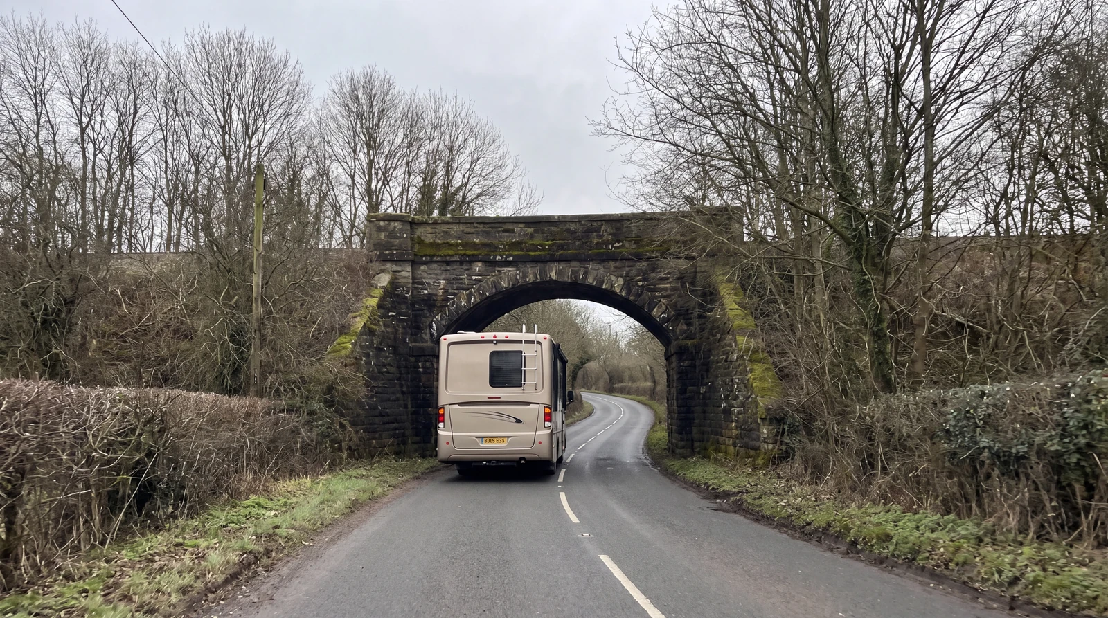 View from behind a Class A motorhome approaching a low stone arch railway bridge over a rural two lane road under overcast light